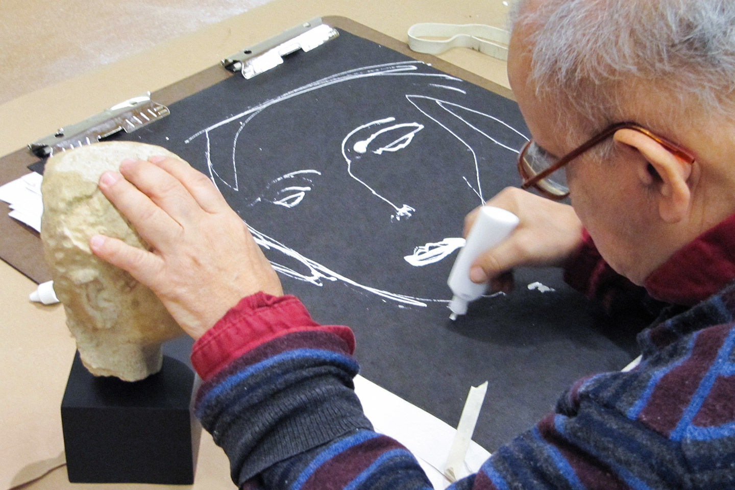 An elderly man wearing glasses draws a faces with his right hand while touching a portrait bust sculpture with his left hand